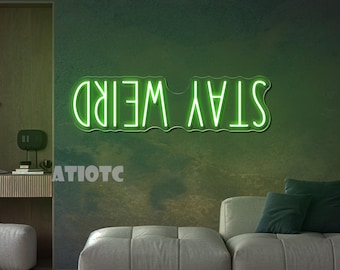 Pode incluir: Uma placa de néon verde que diz "STAY WEIRD" em uma fonte moderna. A placa está montada em uma parede verde escura texturizada. A imagem também mostra uma sala de estar moderna com um sofá cinza e almofadas verdes.