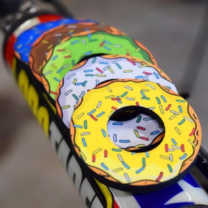May include: A set of colorful donut-shaped bike frame protectors. Each protector features a different frosting color: yellow, white, green, and brown, all with colorful sprinkles. The protectors are designed to fit around a bike frame.