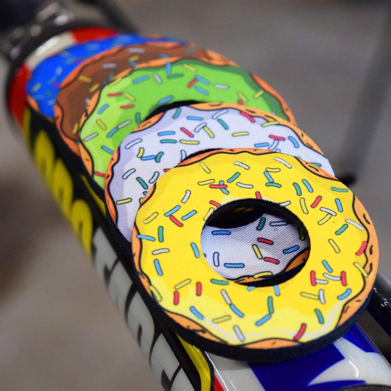 May include: A set of colorful donut-shaped bike frame protectors. Each protector features a different frosting color: yellow, white, green, and brown, all with colorful sprinkles. The protectors are designed to fit around a bike frame.