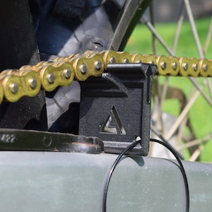May include: A close-up of a motorcycle chain with a black chain guide. The chain is gold-colored and runs through the guide, which has a triangular cutout. The background includes a portion of the motorcycle's wheel and frame.