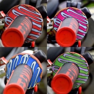 May include: Close-up of four motorcycle handlebar grips with donut-themed designs. Each grip features a different color scheme: red, pink, blue, and green, with white and gray icing details. The grips have a red end cap and a textured gray handle.