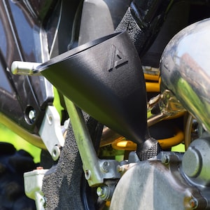 May include: A black funnel with a textured surface and a triangular logo is positioned over a motorcycle engine. The funnel is designed to aid in the pouring of fluids. The motorcycle has silver and yellow components.