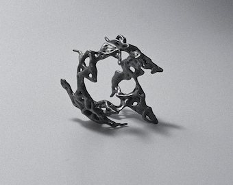 May include: A dark grey ring with an irregular, open-work structure resembling a porous or molten metal texture. The band features multiple jagged protrusions and hollow spaces throughout its circular form, creating a complex, organic, and uneven silhouette.