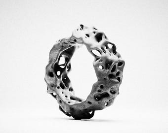 May include: A grey ring with a porous, uneven surface texture featuring numerous irregular holes and organic, crater-like indentations throughout the entire band, creating a complex, open-work structure with a matte finish and a monochromatic appearance.