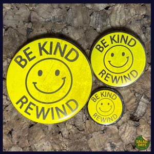 May include: Three yellow buttons with a smiley face design and the words "BE KIND REWIND" in black. The buttons are in different sizes, with a large, medium, and small button.