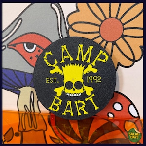May include: A round, black patch with yellow text and a skull design. The text reads "CAMP BART EST. 1992." The patch is set against a background of colorful, cartoonish illustrations, including flowers and a mushroom.