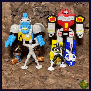 May include: A collection of colorful action figures, including a large blue and gray monster, a red and white robot, and two smaller figures. The figures are set against a textured brown background. The image includes the text "SALAD DAYS".