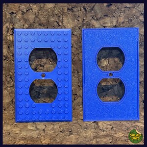 May include: Two blue electrical outlet covers. One cover has a textured surface with circular bumps, and the other has a smooth surface. Both covers have two circular openings for the outlets. The logo "SALAD DAYS" is in the bottom right corner.