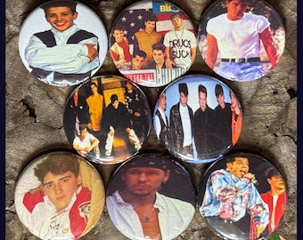 May include: A collection of nine round pinback buttons featuring images of pop stars and bands. The buttons showcase various portraits and group shots, with some including text like "Drugs Suck" and "Salad Days". The buttons are arranged on a dark, textured surface.