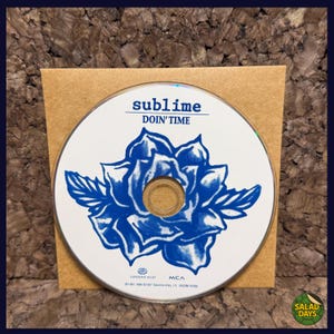 May include: A CD with the text "sublime DOIN' TIME" in blue, featuring a blue floral design on a white background. The CD is in a brown paper sleeve, set against a textured background.