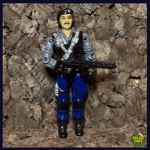 May include: A vintage action figure of a soldier in a black vest, gray sleeves, and blue pants, holding a black machine gun. The figure has a mustache and a black cap. The background is a textured, brown surface.