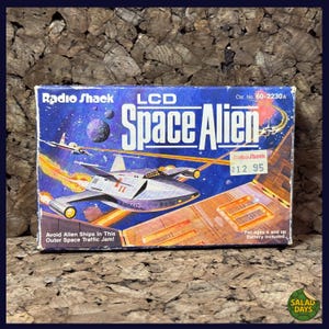 May include: A vintage Radio Shack LCD Space Alien game box. The box features a space scene with a spaceship, planets, and the text "Space Alien." The box also includes the text "Avoid Alien Ships In This Outer Space Traffic Jam!" and the price.