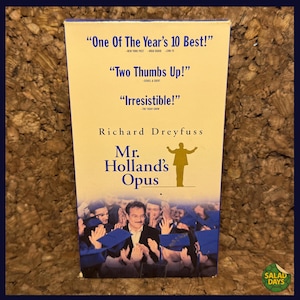 May include: A VHS tape for the movie "Mr. Holland's Opus" featuring Richard Dreyfuss. The cover includes quotes like "One Of The Year's 10 Best!" and "Two Thumbs Up!" with a picture of the cast.