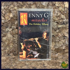 May include: A vintage cassette tape for "ENNY G miracles The Holiday Album." The cover features a musician playing a wind instrument, with text in red and yellow. The cassette is in a clear plastic case.