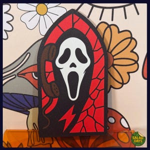 May include: A patch featuring the Ghostface mask from the Scream movie franchise, designed in a stained-glass style. The patch has a red border, black background, and a white mask holding a brown telephone. A red lightning bolt is also visible.