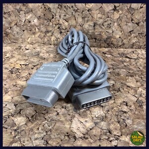 May include: A gray video game cable with two connectors and a coiled wire. The connectors are rectangular with a series of pins. The cable is tied with a black band. The background is a brown, textured surface.