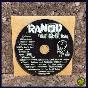 May include: A Rancid "Life Won't Wait" CD album on a brown paper sleeve. The black CD features the band's name and album title, along with a skull graphic and a list of song titles. The CD is set against a dark background.