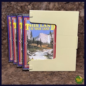 May include: A collection of cassette tapes in a beige case. The tapes have a red, blue, and yellow design with the text "THIS LAND IS YOUR LAND" and a landscape image. The case is open, revealing the tapes.