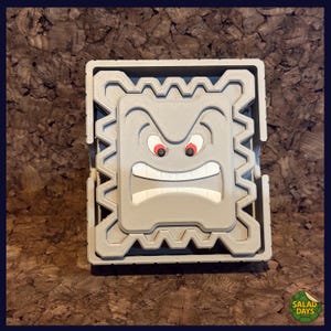 May include: A gray, 3D-printed Thwomp from the Super Mario video game series. The Thwomp has a square shape with a jagged border and a menacing face with red eyes and a wide, toothy grin. The item is sitting on a brown surface.