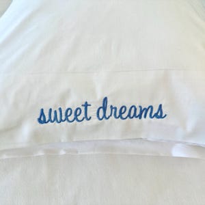 May include: A white pillowcase featuring the words sweet dreams embroidered in blue cursive script. The text is centered on the lower portion of the pillowcase, which has a smooth fabric texture and a clean, simple design for bedding.