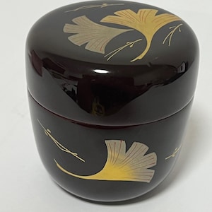 May include: A dark brown, lacquered, round container with a lid. The container is decorated with gold and tan ginkgo leaf designs. The container has a glossy finish and a smooth surface. The container is likely used for storage.