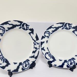 May include: Two white ceramic plates with a navy blue patterned border. The border design features a repeating pattern of abstract shapes and circular motifs. The plates are displayed on black stands, suitable for decorative or dining use.