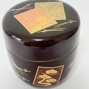 May include: A dark brown cylindrical lidded container with a glossy finish. The lid features a square panel with a pink floral design and a gold bamboo motif. The base displays a rectangular panel with a gold pine tree design on a brown background.