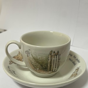 May include: Cream-coloured teacup and saucer set with rabbit and garden illustrations. The cup has a curved handle, and the saucer has text around the edge. The set is made of ceramic.