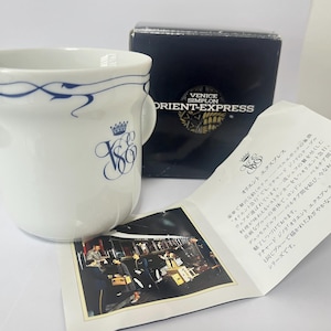 Puede incluir: Una taza de cerámica blanca con un patrón de líneas azules entrelazadas cerca del borde y el monograma VSOE en azul con un logotipo de corona. La taza viene con una caja negra etiquetada Venice Simplon Orient-Express y un folleto informativo.