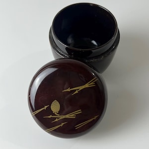May include: A dark brown, lacquered container with a lid. The lid features a gold-coloured pinecone and branch design. The container's interior is black, and the lid is slightly ajar, revealing the inside.