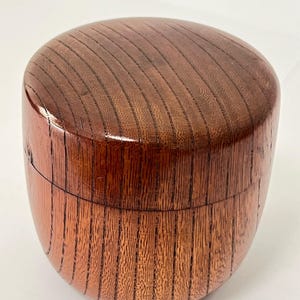 May include: A cylindrical wooden container with a smooth, polished finish and a visible vertical grain pattern. The object features a rounded top lid that fits securely onto the base, creating a seamless appearance with a rich, warm brown color.