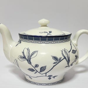 May include: Cream-colored ceramic teapot with a lid and handle. The teapot features a blue floral design and a blue geometric border around the rim and lid. The spout and handle are curved. A small knob sits atop the lid.