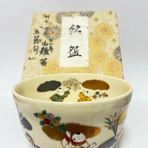 May include: A cream-colored ceramic bowl with a crackle glaze finish, featuring hand-painted illustrations of children in colorful traditional clothing holding floral branches, accented with gold leaf clouds, positioned in front of a matching patterned box.