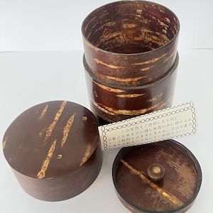 Puede incluir: Un bote de té cilíndrico hecho de corteza de cerezo con un patrón de grano de madera natural. El conjunto incluye un recipiente principal, una tapa y una tapa interior. Se incluye una pequeña etiqueta de papel con texto en japonés.