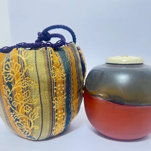 May include: A small, round, fabric pouch with a drawstring closure, featuring a striped pattern in gold, blue, and orange. Next to it is a two-toned ceramic jar, with a red base and a gray top with a cream-colored lid.