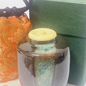Pode incluir: Um pequeno pote de cerâmica marrom com tampa amarela, com esmalte turquesa e marrom. Uma bolsa laranja estampada e uma caixa verde estão no fundo. O pote tem um acabamento brilhante e uma forma arredondada.
