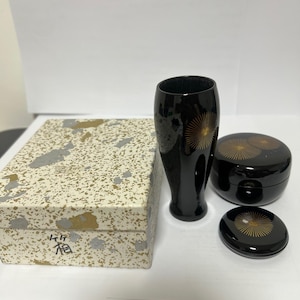 May include: A black lacquerware set comprising a tall vase, a lidded container, and a small dish, all decorated with gold sunburst motifs. A rectangular box with a speckled pattern is also present. The set is likely of Japanese origin.