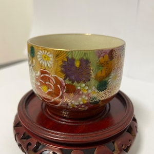 Puede incluir: Un cuenco de cerámica decorativo con un diseño floral en rojo, amarillo, morado y blanco, colocado sobre un soporte de madera rojo oscuro. El cuenco tiene un borde dorado y probablemente se utiliza para exhibición o servicio.