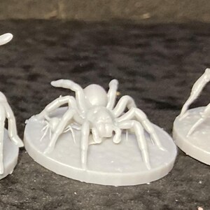 May include: A light gray spider figurine with eight legs and a rounded body, mounted on a circular base. The spider is in a crawling position, with its legs spread out. The figurine is likely made of plastic or resin.