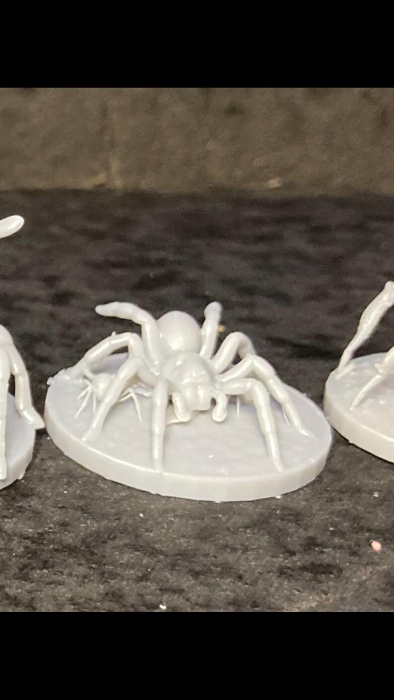 May include: A light gray spider figurine with eight legs and a rounded body, mounted on a circular base. The spider is in a crawling position, with its legs spread out. The figurine is likely made of plastic or resin.