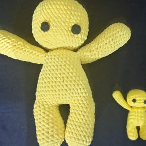 Puede incluir: Dos muñecos amarillos de ganchillo con los brazos extendidos y ojos de botón negros. Un muñeco es significativamente más grande que el otro. Los muñecos están hechos a mano y tienen un aspecto suave y texturizado. El fondo es azul oscuro.
