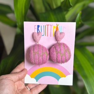 May include: Pink felt earrings with heart-shaped tops and round, textured bottoms. The earrings are displayed on a pink card with the text "FRUITY PUNK" above a rainbow graphic. The background features green foliage.