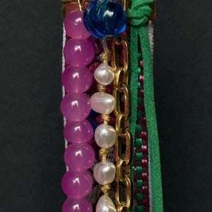 Macro shot highlighting pink and pearl bead strands with gold chain and green cord over decorative embroidered ribbon.
