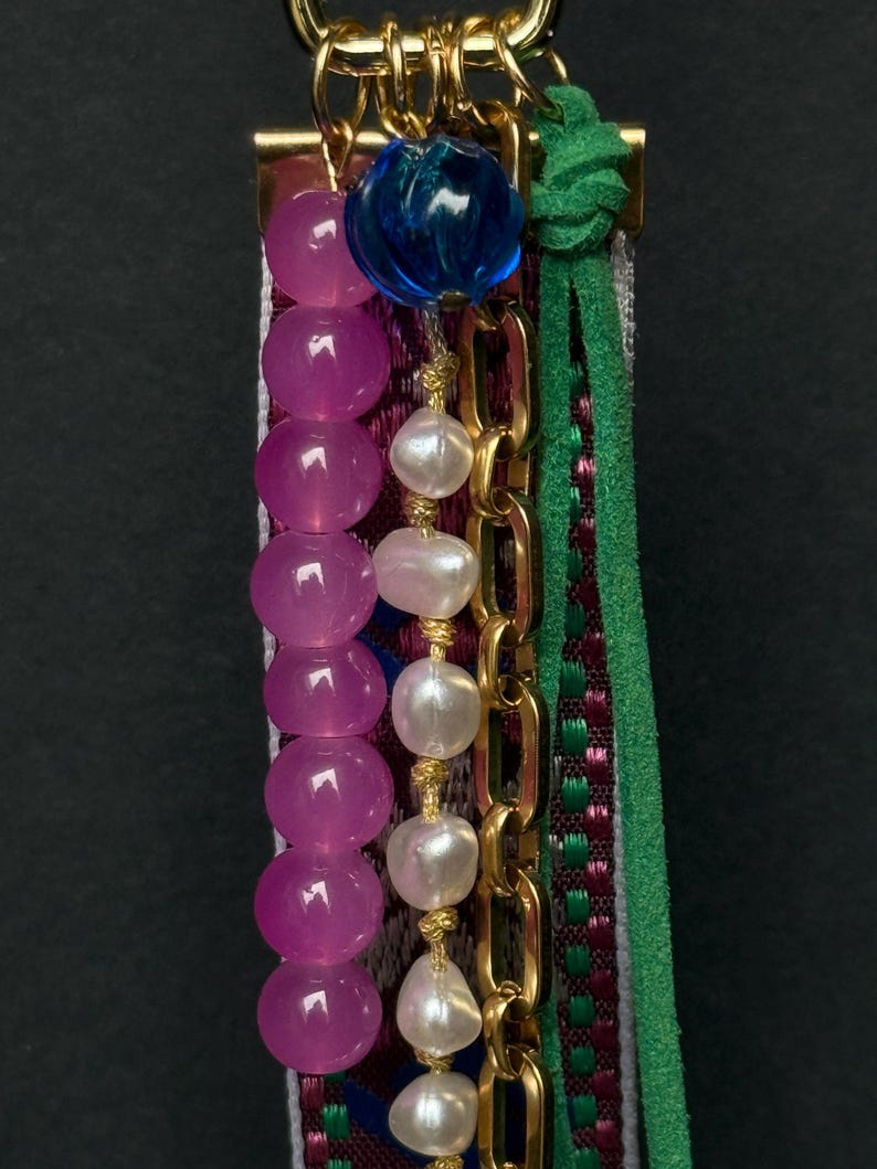 Macro shot highlighting pink and pearl bead strands with gold chain and green cord over decorative embroidered ribbon.