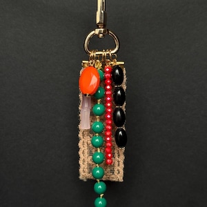 Handmade bag charm with gold clasp, beige lace ribbon, and strands of orange, green, red, pink, and black beads in various shapes and finishes.