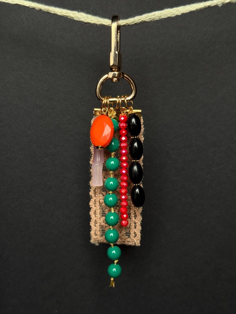 Handmade bag charm with gold clasp, beige lace ribbon, and strands of orange, green, red, pink, and black beads in various shapes and finishes.