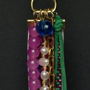 Close-up of top section showing blue glass bead, gold rings, and strands of pink and pearl beads next to green suede cord.