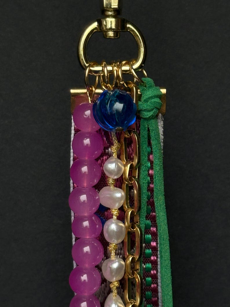 Close-up of top section showing blue glass bead, gold rings, and strands of pink and pearl beads next to green suede cord.
