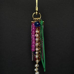 Op de afbeelding: Een handgemaakte sleutelhanger met een goudkleurige sluiting. De sleutelhanger heeft een mix van kralen, waaronder roze en witte parels, een gouden ketting en een groene suède kwast. Een kleurrijke geweven stoffen strook voegt details toe.