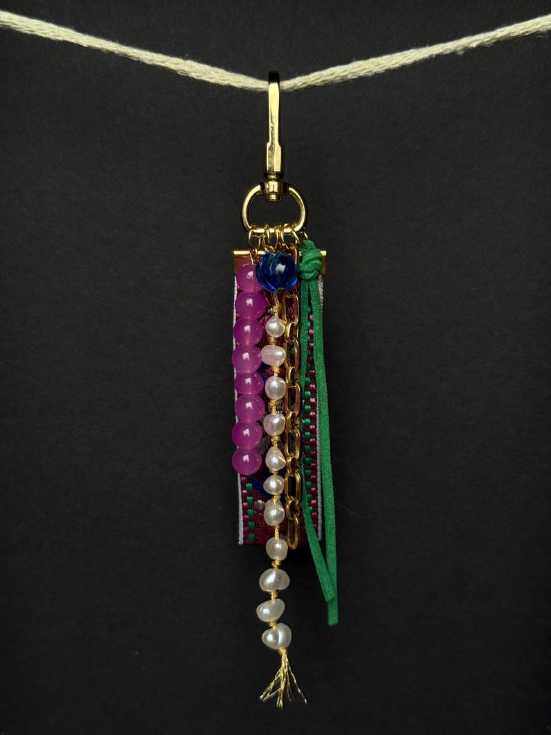 Handmade bag charm with gold clasp, fuchsia and pearl bead strands, green suede cord, and blue glass bead on an embroidered ribbon with pink and green details.